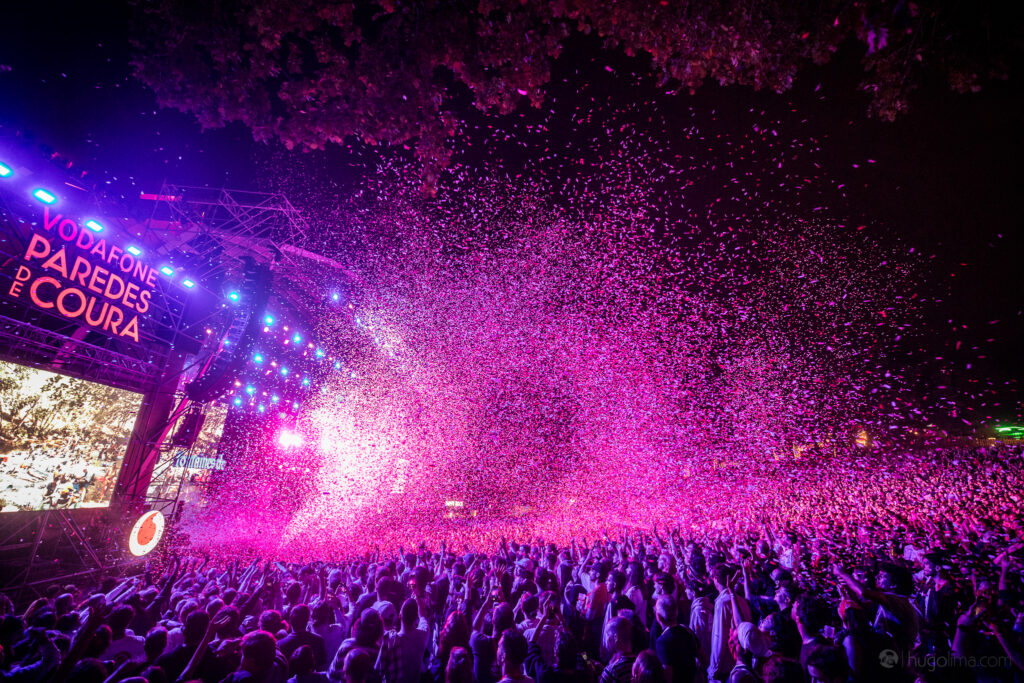 A picture of the main stage of the Paredes de Coura festival at night, with confetti being thrown over the crowd under a purple-ish light
